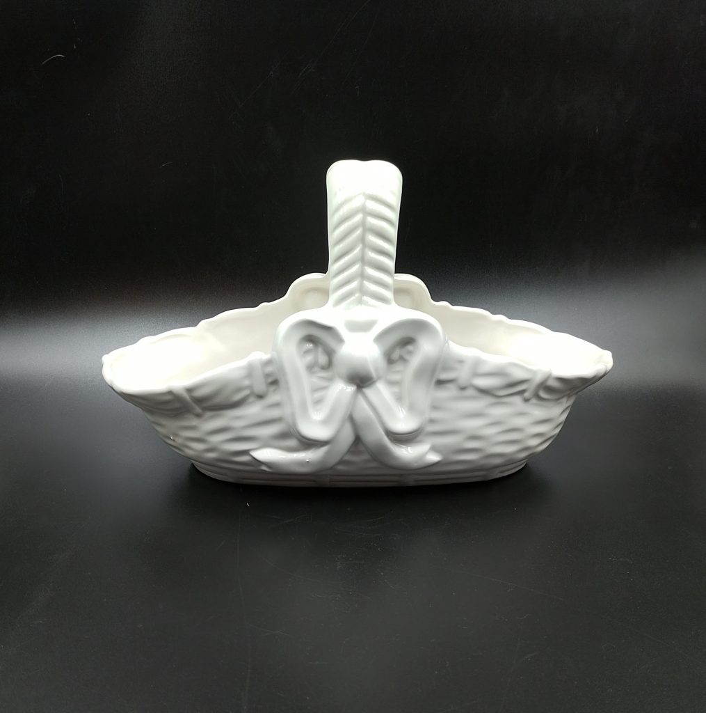 Biały ceramiczny koszyk na czarnym tle, koszyk posiada tłoczone zdobienia imitujące splot kosza oraz tłoczoną kokardę